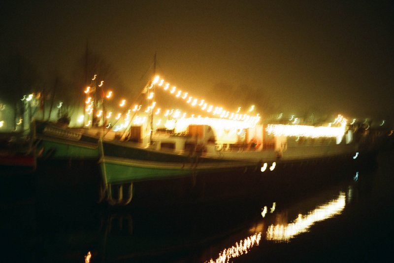 The image captures a serene night scene at a harbor. The central focus is a boat docking at a well-lit pier. Alongside the dock, there's a collection of boats and docks visible, adding to the maritime atmosphere. There are various lights illuminating the area, creating a contrast with the surrounding darkness. The lighting suggests a vibrant and active harbor, while the calm water reflects the illumination.