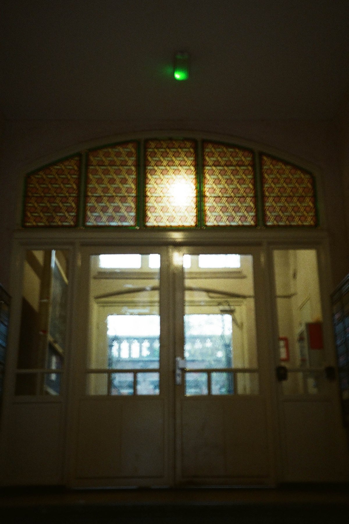  The image features an interior entrance way of a building during nighttime. There is a doorway with a visible light source above it, illuminating the area immediately around it. On either side of the doorway are large stained glass windows adorned with intricate patterns, predominantly in shades of blue and red, which suggest a classical or historic architectural style. The walls surrounding the entranceway are painted in a darker shade, possibly black or navy blue, providing a stark contrast to the lighter ceiling. 