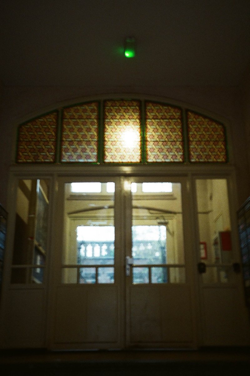 The image features an interior entrance way of a building during nighttime. There is a doorway with a visible light source above it, illuminating the area immediately around it. On either side of the doorway are large stained glass windows adorned with intricate patterns, predominantly in shades of blue and red, which suggest a classical or historic architectural style. The walls surrounding the entranceway are painted in a darker shade, possibly black or navy blue, providing a stark contrast to the lighter ceiling.