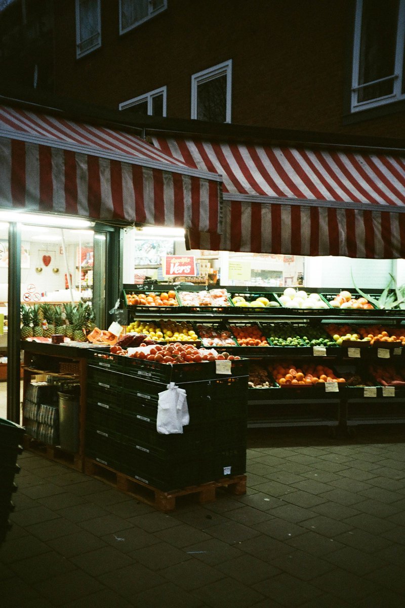 The image depicts an outdoor fruit and vegetable market at night. There are several items of produce visible under various umbrellas, with the majority being apples. The fruits are arranged in bins and on shelves for display. A sign advertising fresh produce is also present. The setting appears to be a city street corner with a brick building in the background.