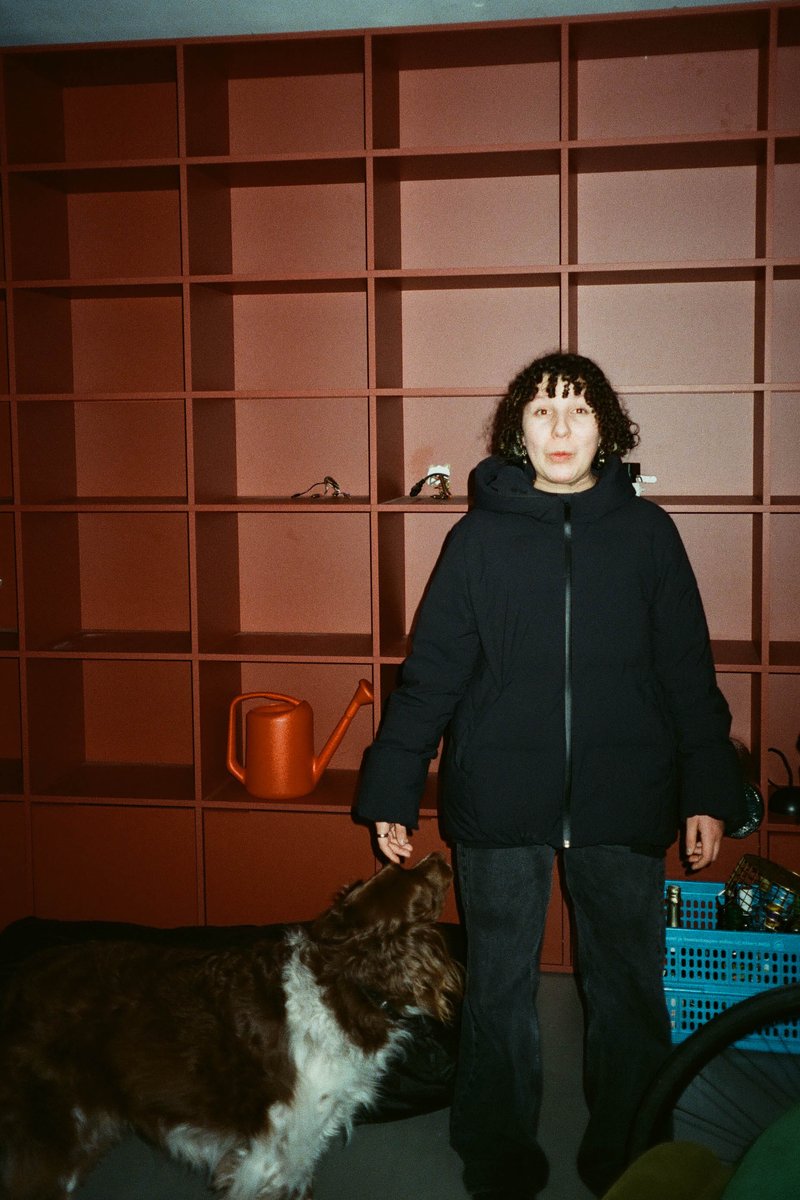 This is a black and white photograph featuring a woman standing next to an array of open bookshelves against a red background. The woman appears to be posing for the photo. She has short hair, is wearing a dark coat, and has a dog on a leash at her feet. The shelves are filled with various items, suggesting a personal collection or a study area. There's also a potted plant nearby.