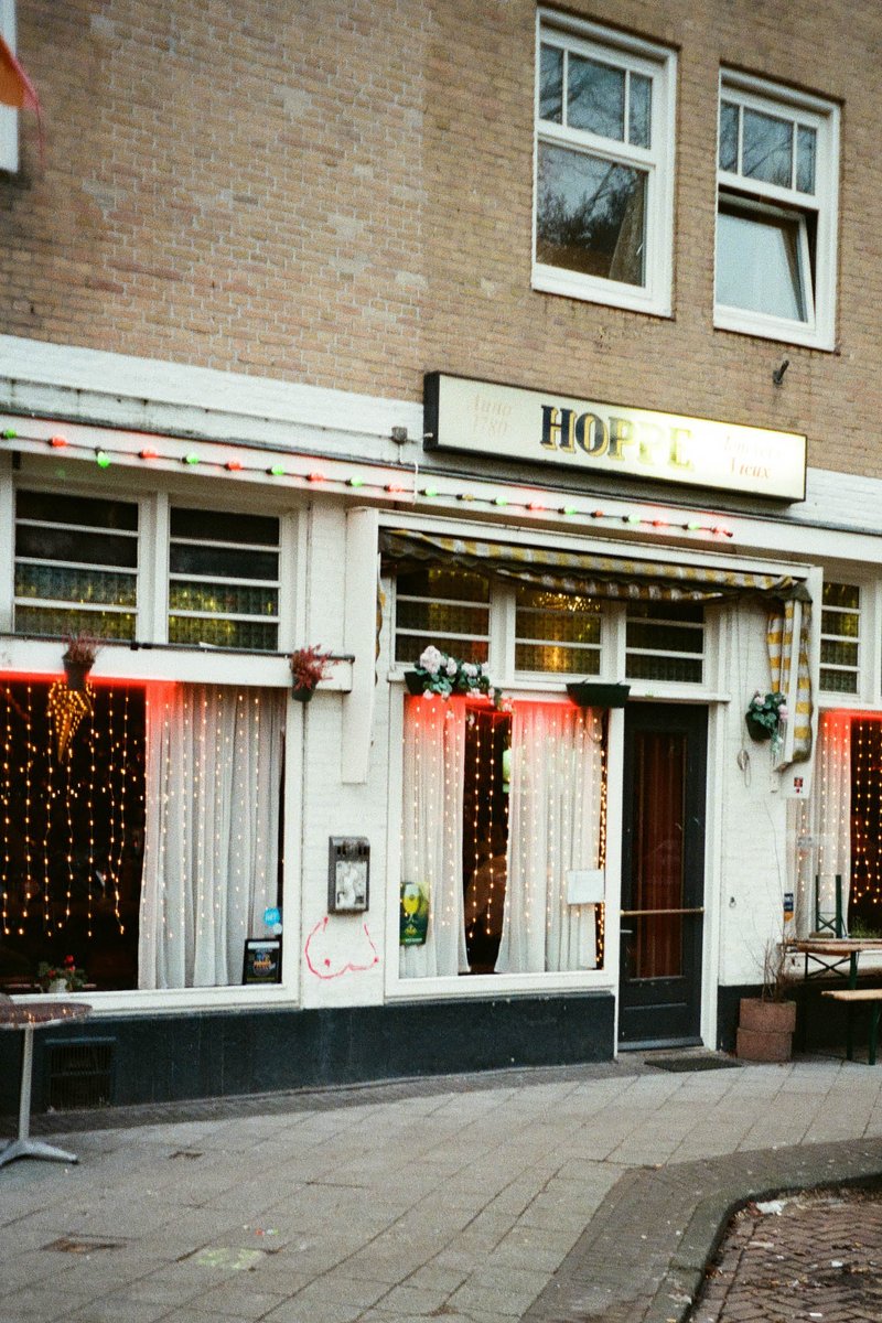 The image shows an exterior view of a building with the name "Hope" visible above its entrance. In front of the building is a festive street scene decorated for Christmas or another holiday, suggesting a festive atmosphere. The decorations include colorful lights and garlands, which create a warm and inviting ambiance. There's no text present in the image to provide additional context.