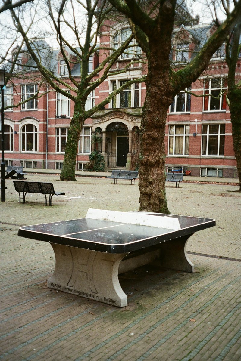 The image features an urban setting with a black table as the main subject. This table is situated in a park-like area, with a bench nearby. In the background, there are buildings with traditional architecture and trees. The park appears to be empty at this time of day.