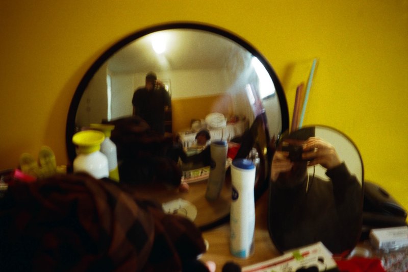 The image shows two mirror reflections: one of a person taking a photo of another individual. The background is indistinct but features warm tones, including a yellow wall and brown items on surfaces like the table. There are also personal care items visible. The setting appears to be an indoor space with various objects around, giving it a lived-in feel.