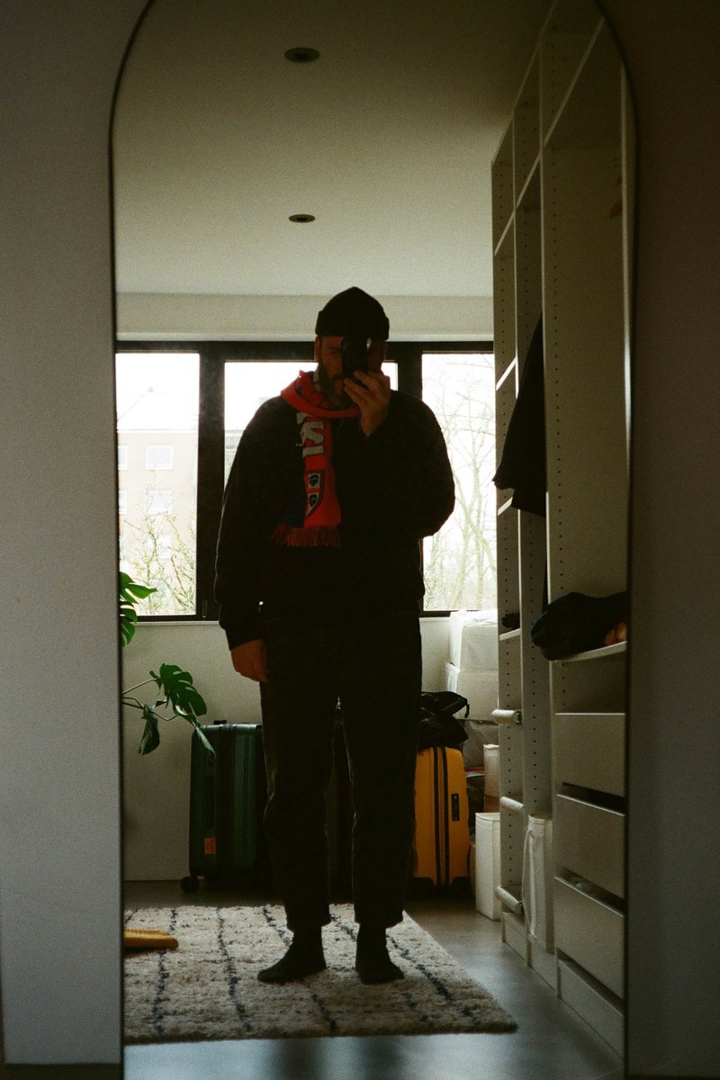 The image is a photograph taken indoors. There is a person standing in a room holding up a camera phone. The individual is wearing dark clothing with a red object in the background. The flooring is light, and there is an open door leading to another area.