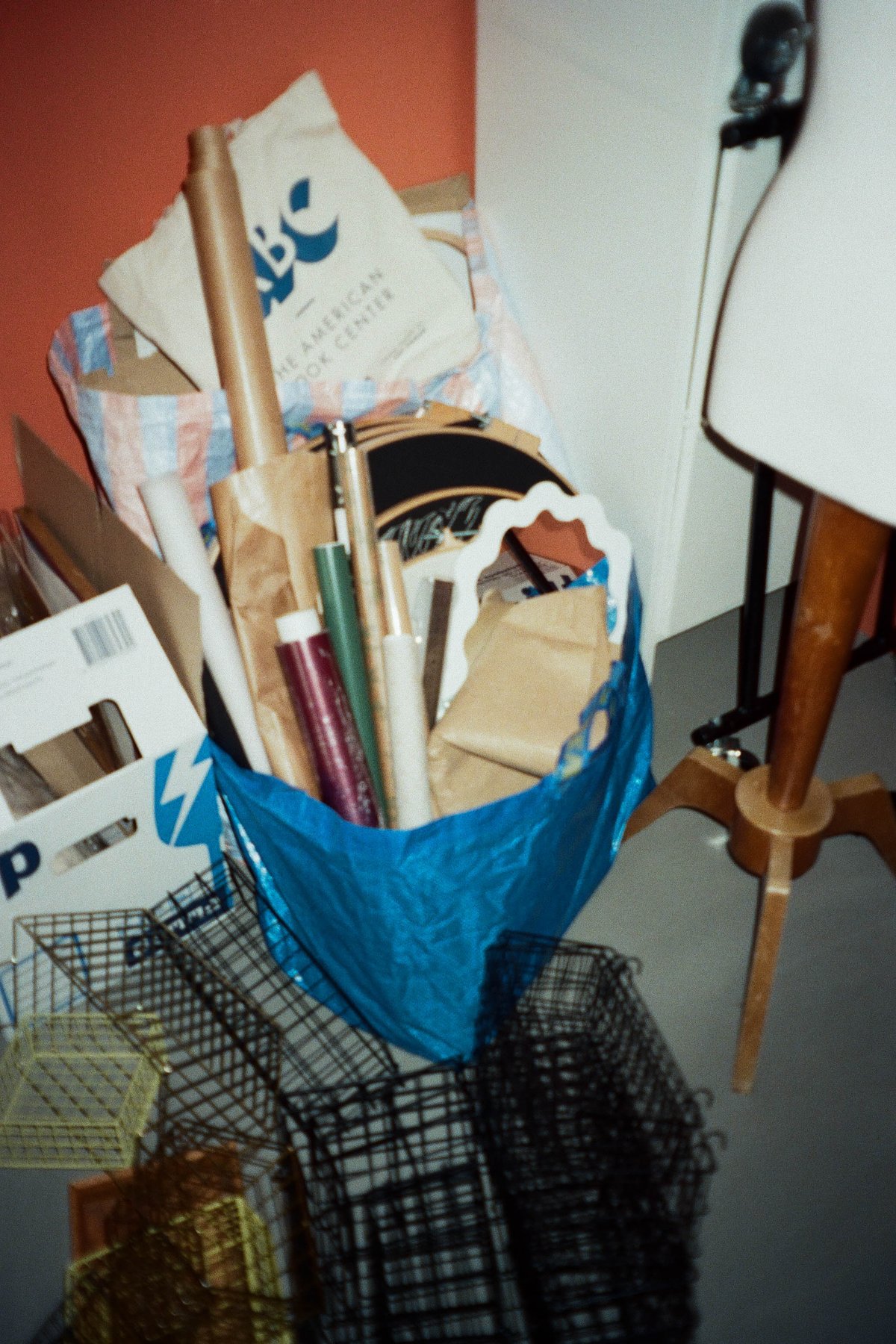  The image depicts a room with various items scattered around. There is a table against one wall, and a blue bag filled with different objects, including what appear to be art supplies, on top of it. Nearby, there are boxes containing more materials, suggesting an ongoing craft or art project. A floor lamp with a visible bulb can also be seen in the room. The overall scene suggests a workspace or creative environment. 
