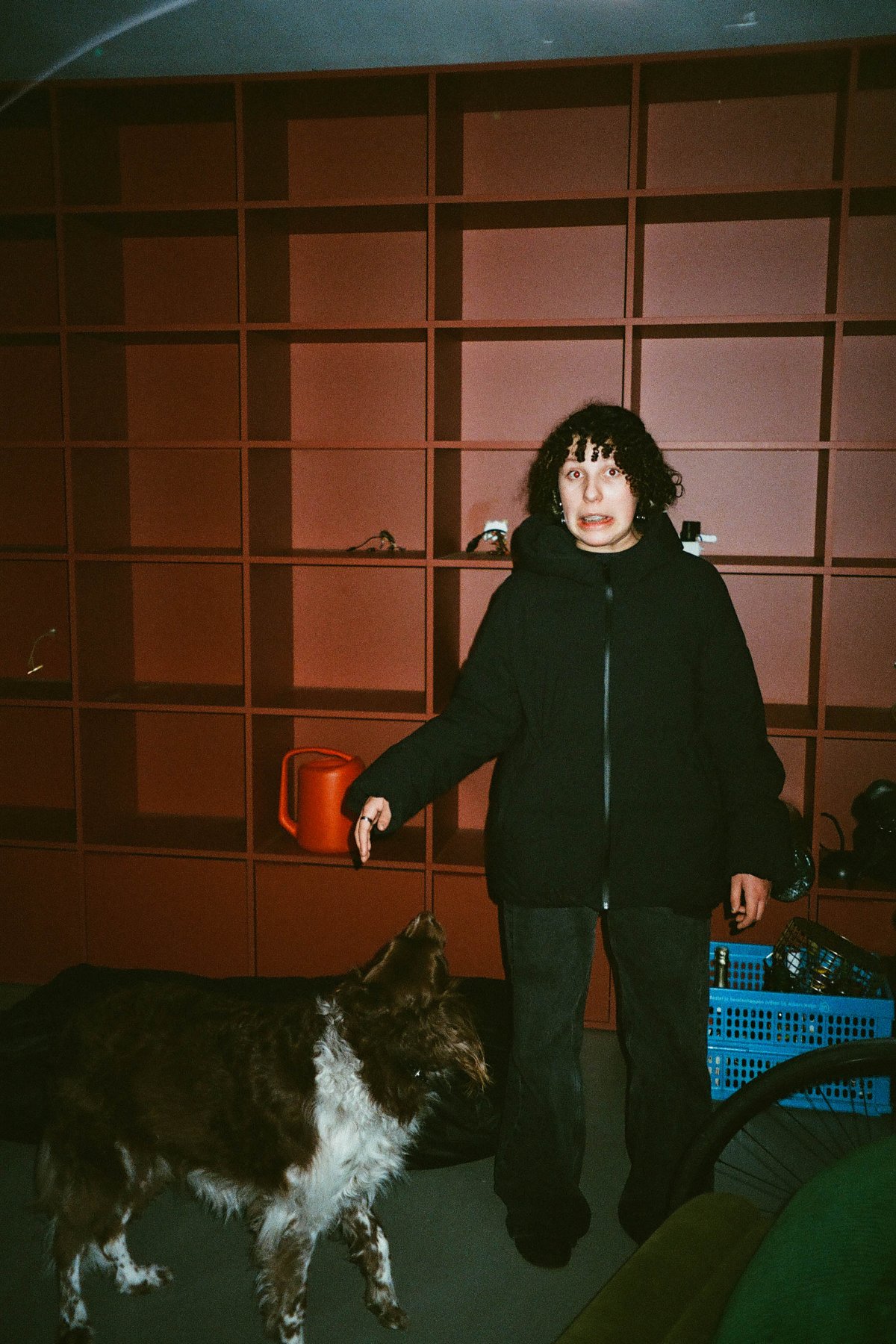  The image is a grainy black and white photograph featuring an individual standing indoors with a dog at their feet. The person appears to be pointing towards something or someone off-camera. In the background, there's a bookshelf filled with numerous items, which suggests this might be a home environment. 