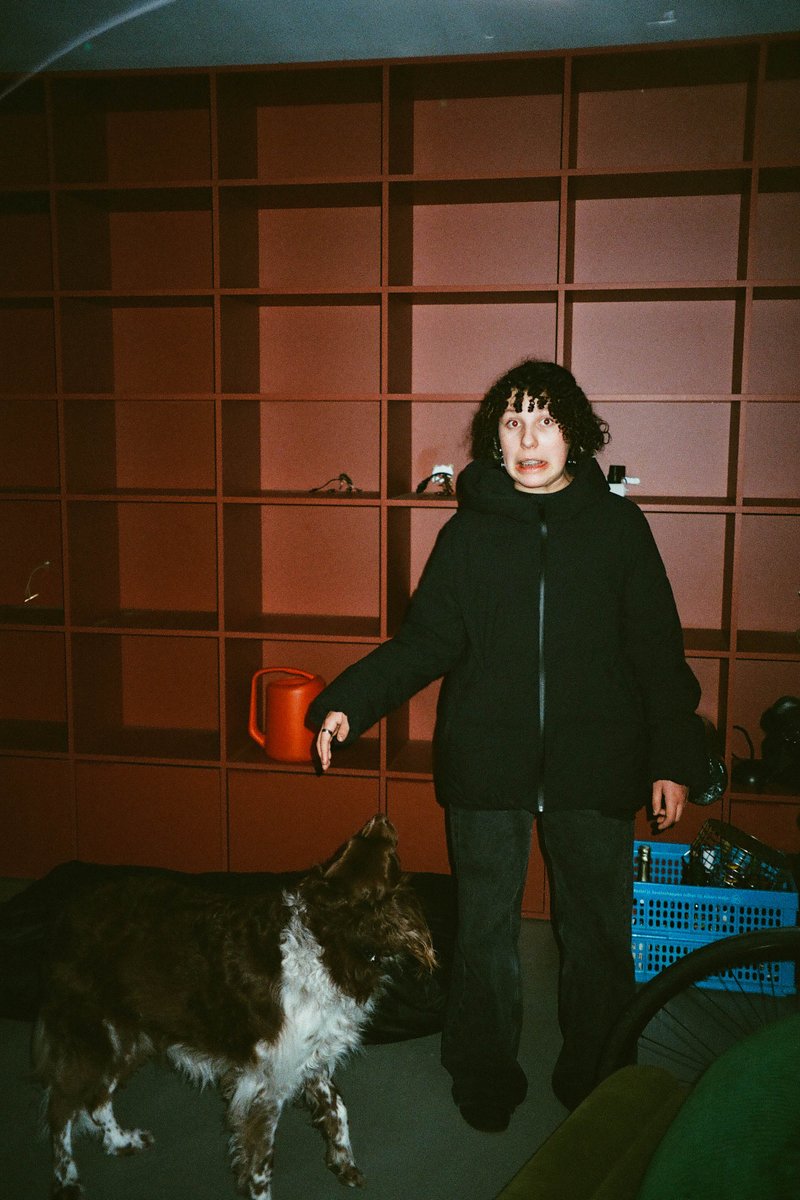 The image is a grainy black and white photograph featuring an individual standing indoors with a dog at their feet. The person appears to be pointing towards something or someone off-camera. In the background, there's a bookshelf filled with numerous items, which suggests this might be a home environment.
