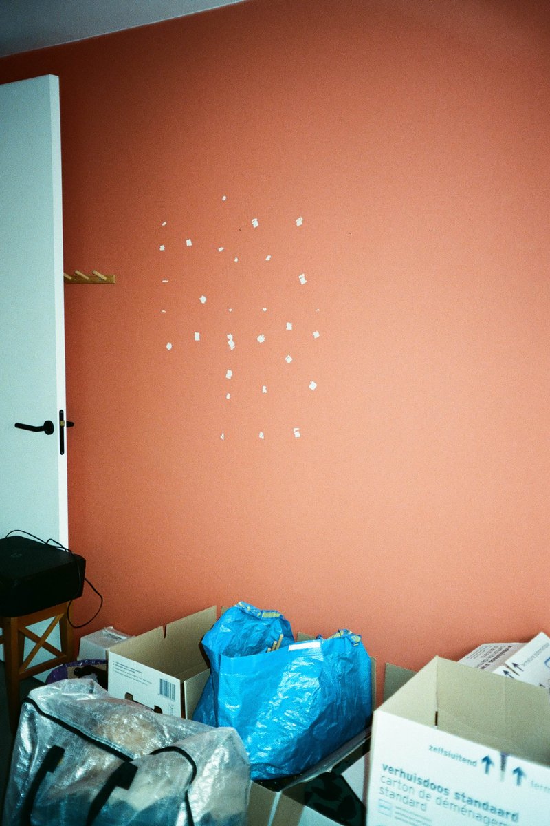 The image shows a room with a white door. On the wall behind the door is a large, rectangular decoration composed of several small dots arranged in a pattern that is not immediately apparent from this description. To the left of the door is a small section of an orange wall. Below the door, there are cardboard boxes and other items scattered on the floor, suggesting the room might be used for storage or work.