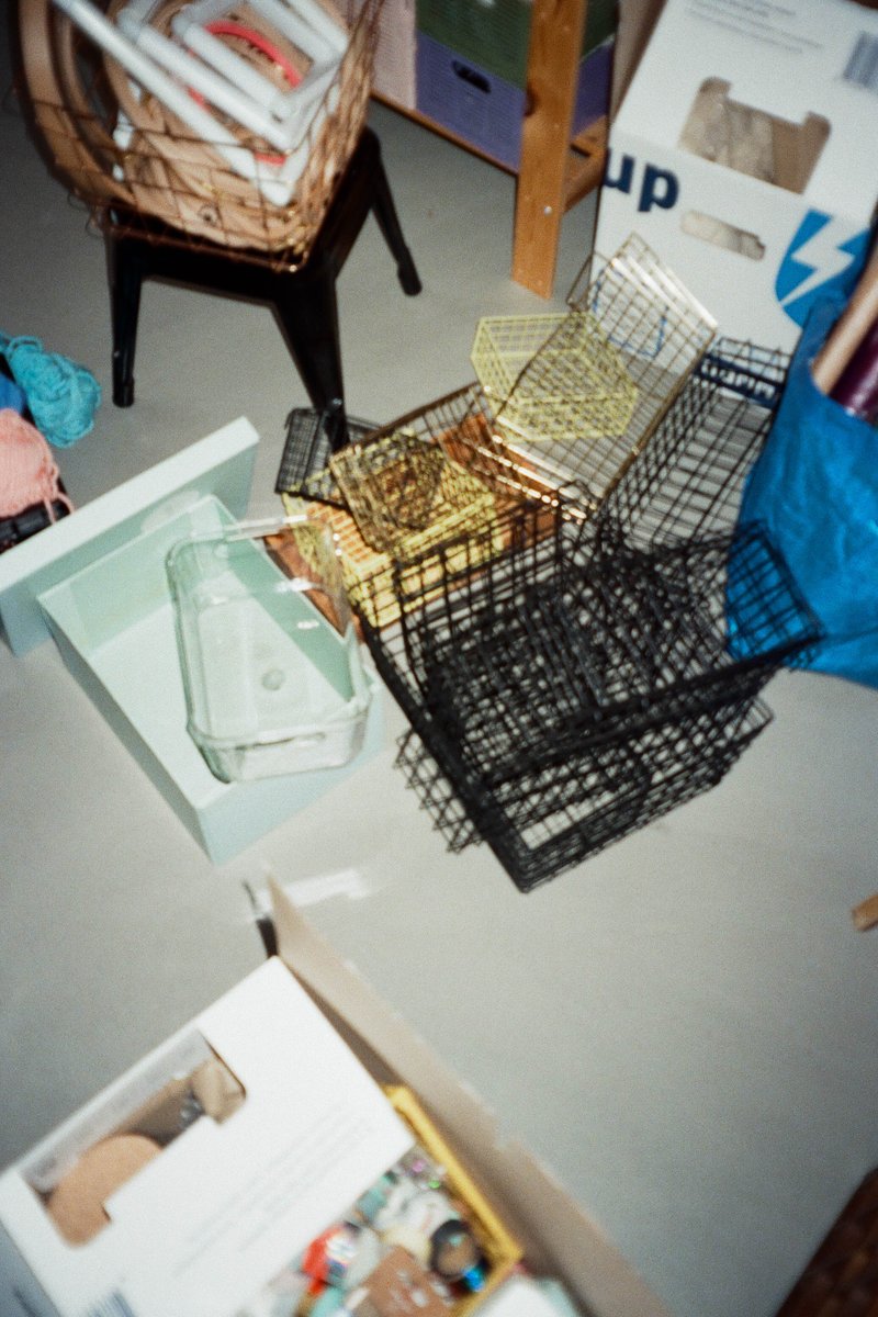 The image shows a cluttered room with several black plastic storage crates stacked haphazardly. There's a pile of what appear to be papers or documents on the floor. To the right, there is a collection of items on the table, including books and boxes that are organized in no particular order. The background reveals additional clutter with cardboard boxes and other household items scattered about, suggesting a storage area or a room in need of tidying up.