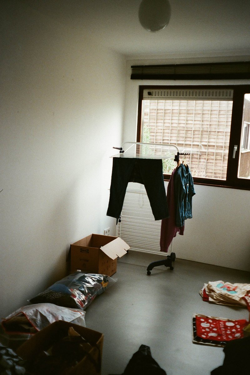 The image shows an interior space that appears to be a room with minimal furniture. On the right side of the room, there is a window with blinds partially open, allowing some natural light into the space. In front of the window, hanging on a clothes hanger, is a shirt and a pair of pants, suggesting someone may have recently arrived or is about to leave. Below these items, there are several cardboard boxes stacked against each other, indicating recent or ongoing packing or unpacking activity. On the left side of the room, there appears to be a small desk with what seems like a computer monitor on it and possibly some papers or books nearby. Above the desk, a shelf holds various objects, including items that might be related to the person living here, such as photos or decorations. The overall impression is that of a lived-in space, perhaps a home or an apartment, with signs of recent activity, possibly due to moving in or out.