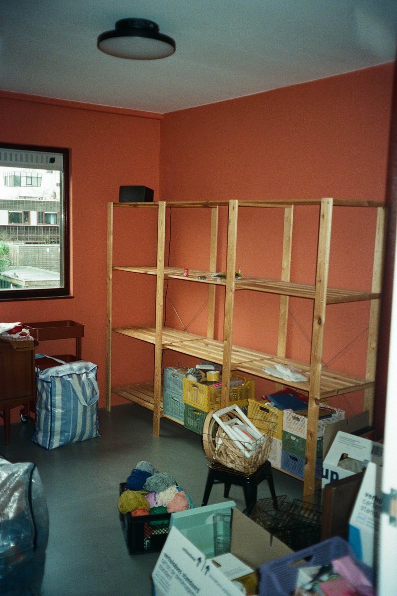 The image shows an indoor space, possibly a room with a red wall. In the center of the room stands a wooden shelving unit that is empty and appears ready for use. To the right, there's a collection of items including various types of containers like buckets and tubs, as well as some boxes, which are grouped together. The presence of these items suggests this could be a storage or organizing area. In the background, there is a window with a curtain partially drawn, allowing natural light into the room. There's no clear indication of any people in the image.