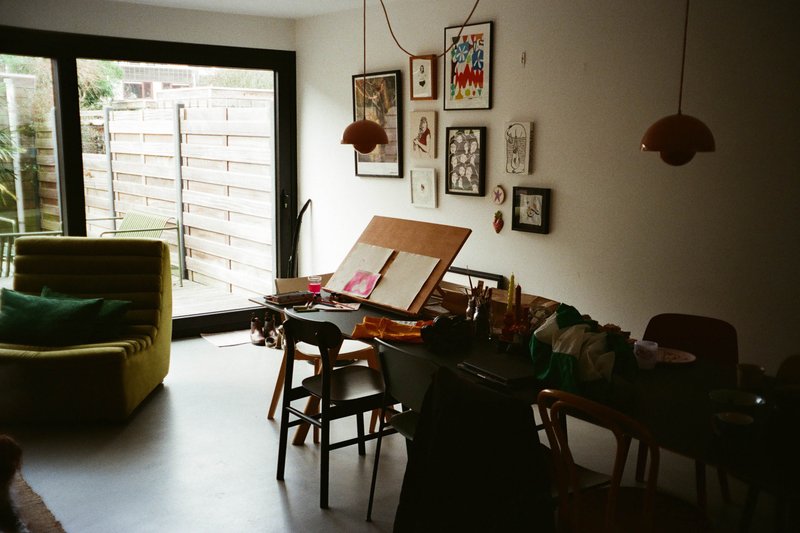 The image shows a spacious room with a modern interior design. Dominating the scene is a white sofa adorned with a colorful throw blanket, positioned against a clean wall painted in a light shade. In front of the couch, there's a wooden table with a few items on top, including what appears to be a small collection of books. Adjacent to this setup, a window lets in natural light, and through it, one can glimpse lush green trees outside, suggesting an urban setting. A painting hangs on the wall, adding a touch of artistry to the space. The room is well-lit and tidy, creating a comfortable and inviting atmosphere.