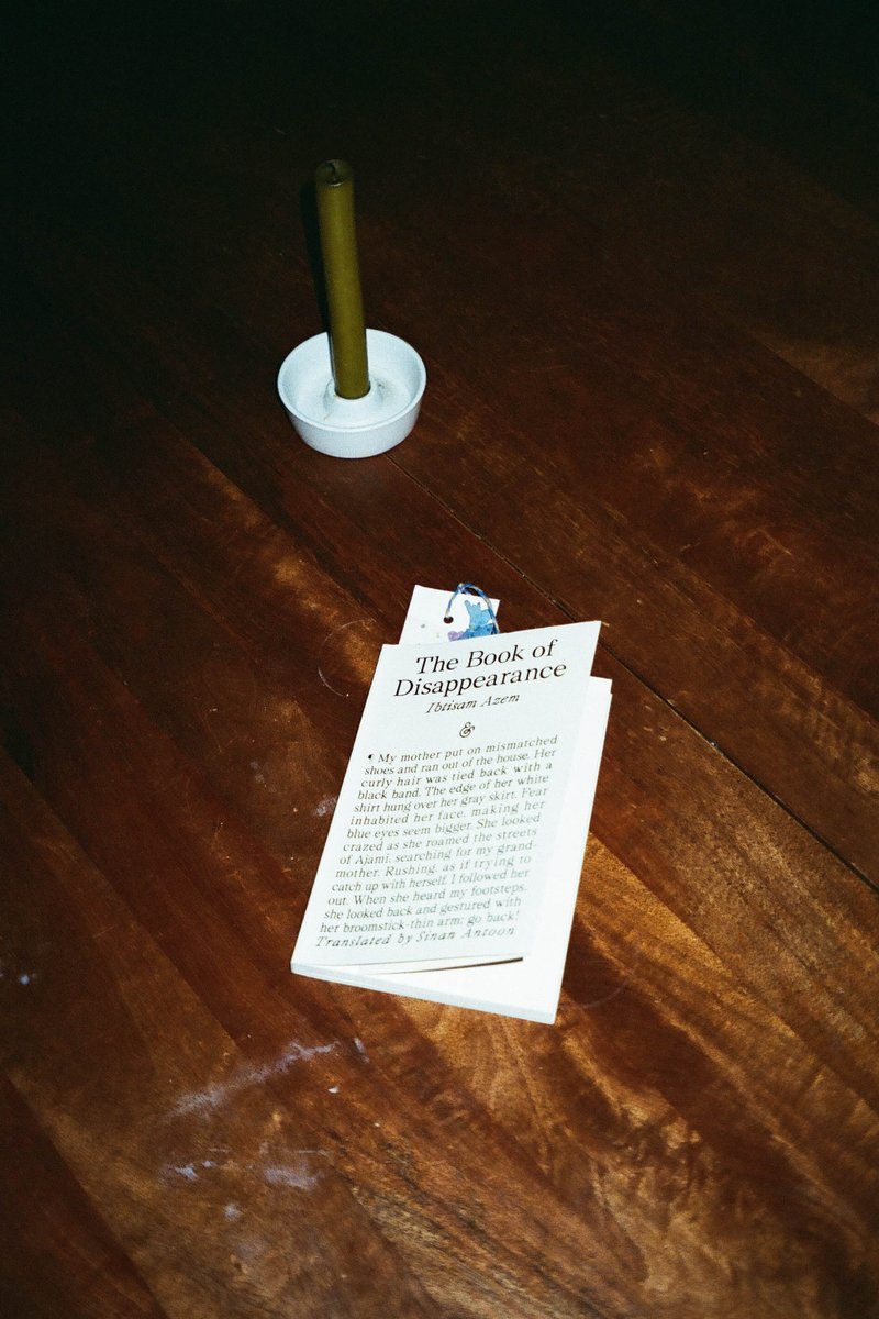 The image depicts a small, dark room with a wooden floor. In the center of the frame, there's a piece of paper that appears to be handwritten. On the left side of the paper, there is a yellow candle. Next to the paper and the candle, there are two white items: one is round and could be a small container or lid, while the other item looks like a thin object that might be part of a toy set. The focus of this image seems to be on the handwritten piece of paper with the yellow candle next to it.