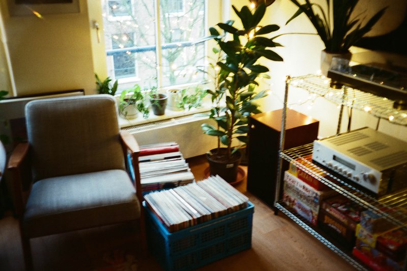 The image depicts a cluttered room with a cozy ambiance. There is a small sitting area featuring a chair and a table holding various items, including books and what appears to be board games. To the right of this space, there's a potted plant adding a touch of greenery. A large window allows natural light to illuminate the room, and outside it, trees can be seen, indicating that this space might be in an apartment or a condo.