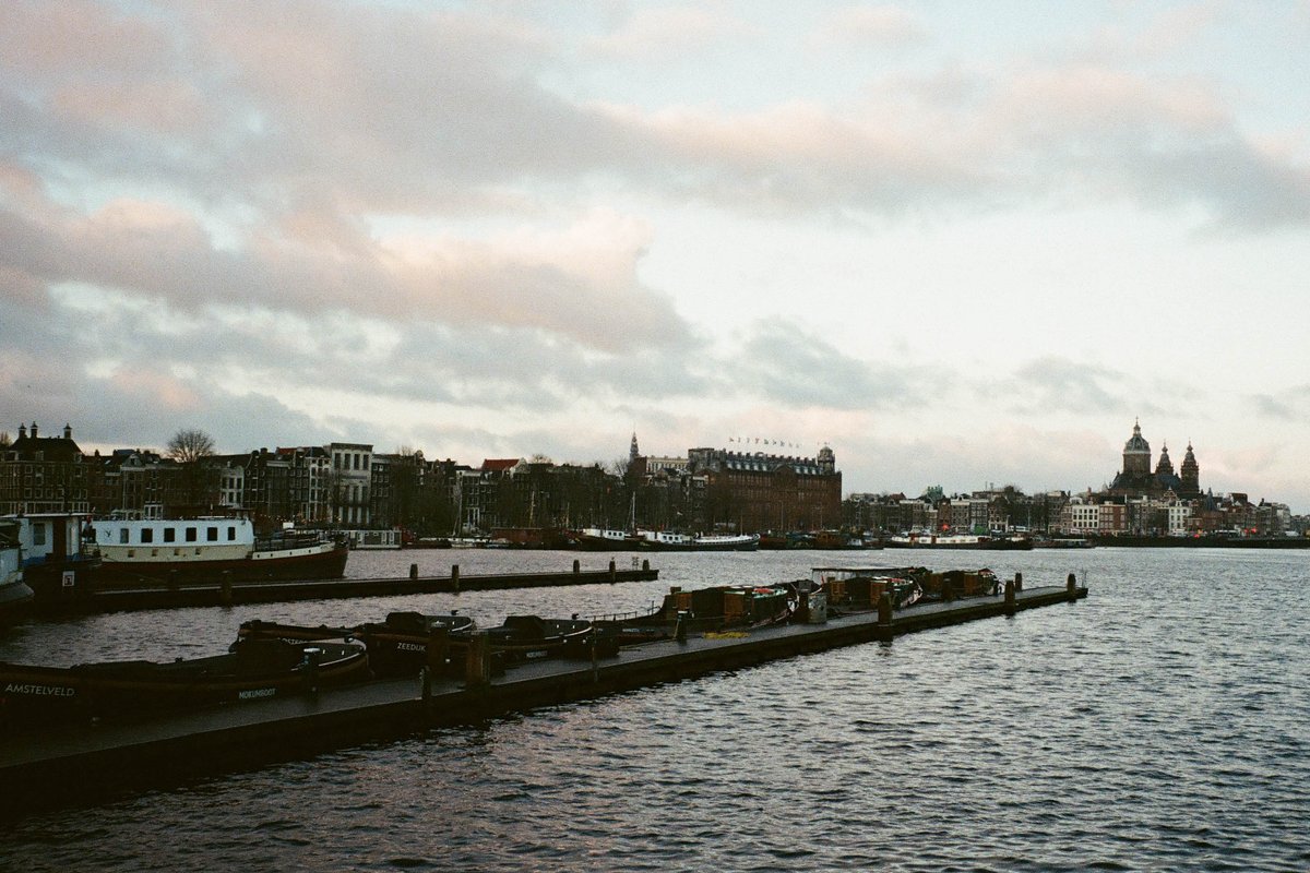  The image depicts a cityscape with a body of water leading to a docked boat. On the left side, there are multiple boats tied up at a pier, and the right side features buildings that appear to be houses or shops, possibly indicating a residential or commercial area. In the background, you can see an overcast sky, suggesting it might be cloudy or raining. The overall scene conveys a peaceful, urban setting with waterfront properties. 