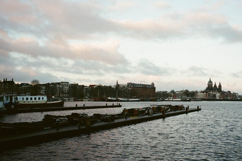 The image depicts a cityscape with a body of water leading to a docked boat. On the left side, there are multiple boats tied up at a pier, and the right side features buildings that appear to be houses or shops, possibly indicating a residential or commercial area. In the background, you can see an overcast sky, suggesting it might be cloudy or raining. The overall scene conveys a peaceful, urban setting with waterfront properties.
