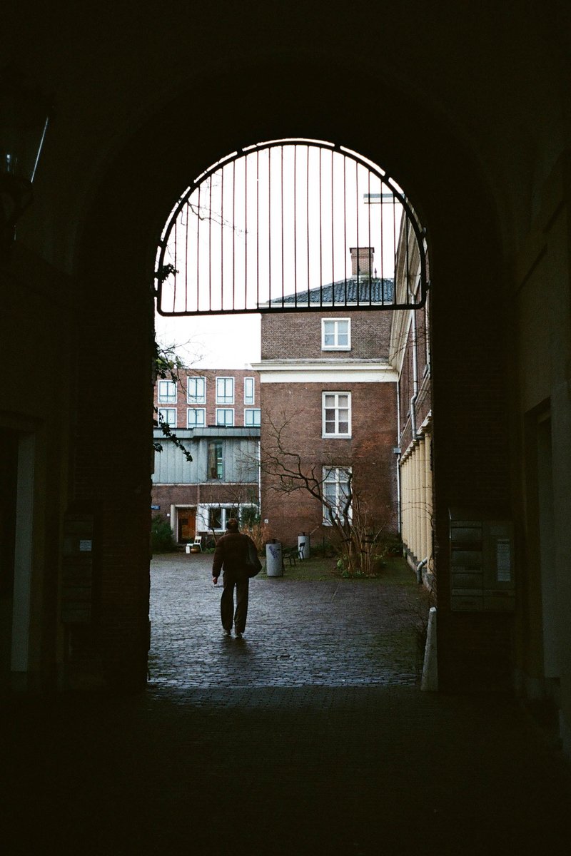 The image shows an architectural scene featuring an arched doorway with bars across it, suggesting security measures. Through the windowed arch, there's a glimpse of what appears to be a person or object in the distance, though details are obscured by the grating. It's a black and white photo that seems to depict an urban setting.