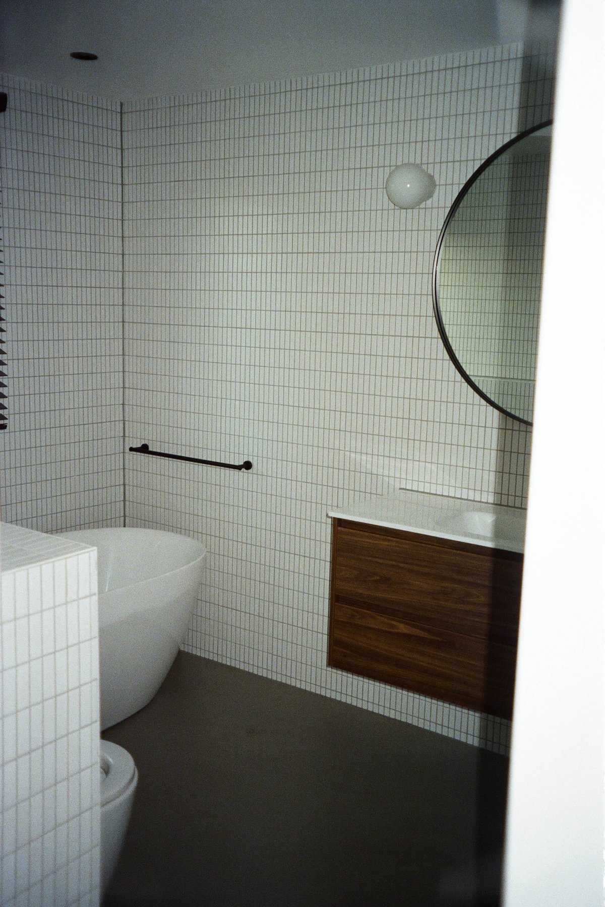  The image showcases a modern bathroom, characterized by its minimalist design and monochromatic color scheme. The room is dominated by a large white tiled floor that contrasts with the pristine white walls. A striking feature of the bathroom is the black and white checkered tiles adorning the back wall above which hangs a sleek, long rectangular mirror, reflecting the clean, uncluttered space within. To the left, a wooden cabinet offers storage, while on the right, a white sink with a simple faucet rests against the wall. Further to the right, a white toilet is tucked neatly into the corner of the room. The floor beneath the toilet bears a darker shade of brown, providing visual interest and breaking up the otherwise uniform palette. A notable detail is the round, black object resting on the sink, adding an element of intrigue to the scene. 