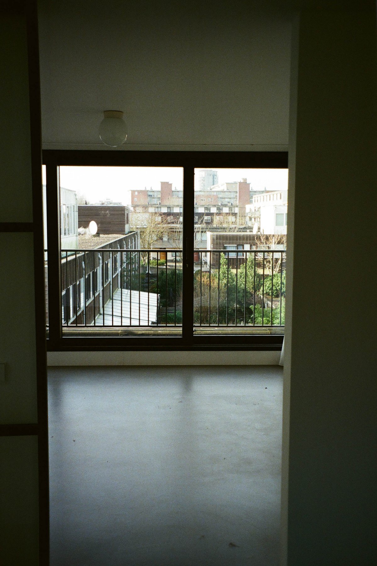  The image shows an interior space with a balcony overlooking a cityscape. There's a large window in the foreground, offering a view of buildings in the distance. The room appears clean and modern, possibly an open concept living area or studio apartment, as indicated by the presence of a dining table that extends to the right edge of the image. The balcony door is visible, suggesting access to outdoor space. There are no texts or people present in the image. 
