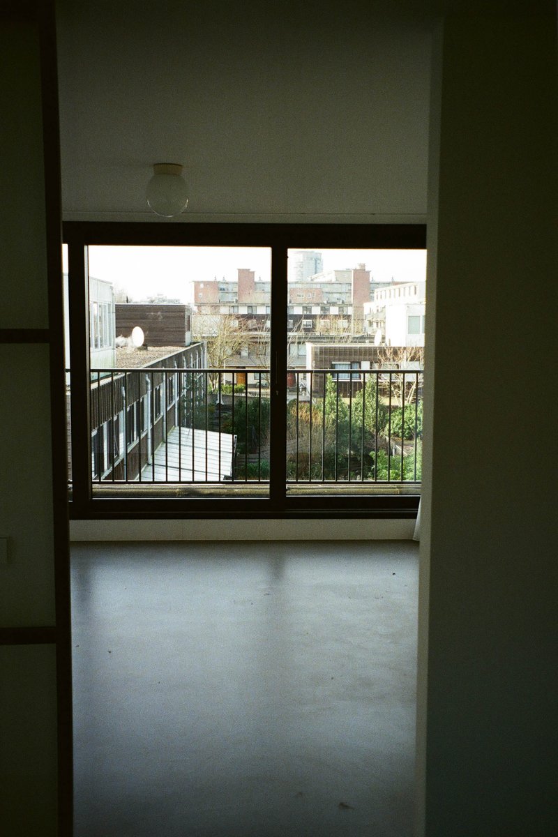 The image shows an interior space with a balcony overlooking a cityscape. There's a large window in the foreground, offering a view of buildings in the distance. The room appears clean and modern, possibly an open concept living area or studio apartment, as indicated by the presence of a dining table that extends to the right edge of the image. The balcony door is visible, suggesting access to outdoor space. There are no texts or people present in the image.