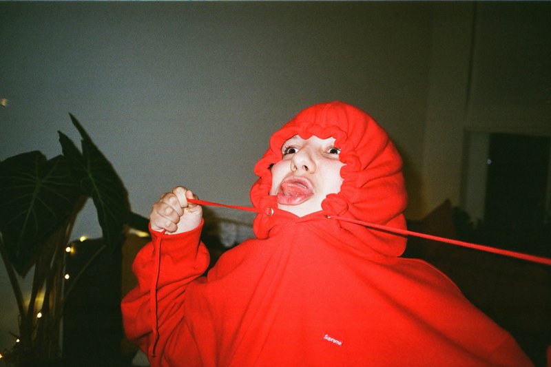 The image features a person with a red hoodie over their head, holding an object that could be a string or ribbon. They are positioned indoors, possibly against a wall adorned with lights or candles. The individual appears to be standing and is the main focus of the photograph. The overall scene suggests a casual indoor setting.