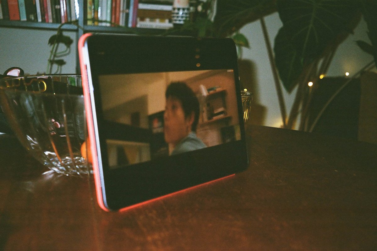  The image shows a small screen displaying a video featuring a man speaking with another person, possibly in a room with a shelf in the background. This device is placed on a table where there's also a bottle and a cup, suggesting a casual indoor setting. 