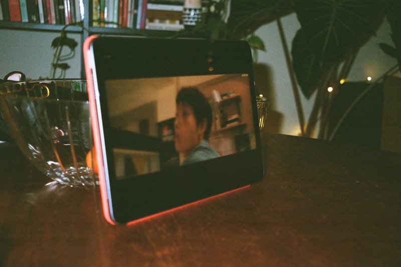 The image shows a small screen displaying a video featuring a man speaking with another person, possibly in a room with a shelf in the background. This device is placed on a table where there's also a bottle and a cup, suggesting a casual indoor setting.