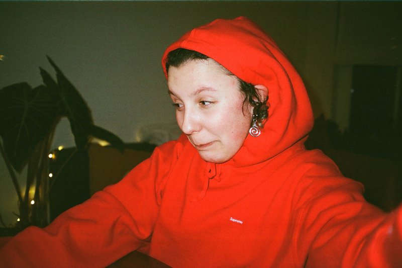 The image shows a person wearing a red hoodie with their hood up, capturing a selfie using a phone or camera. They appear to be indoors, possibly at night given the artificial lighting visible. The person has short hair and is gazing off to the side, away from the camera.