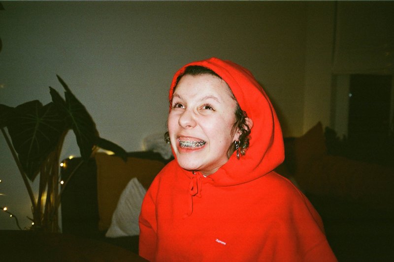 In the image, there is a young woman standing indoors. She is wearing a red hoodie and has a wide smile on her face. Her hair is brown, and she seems to be looking at something off-camera with joy. The background of the photo is simple and contains a plant to one side and a couch on the other.