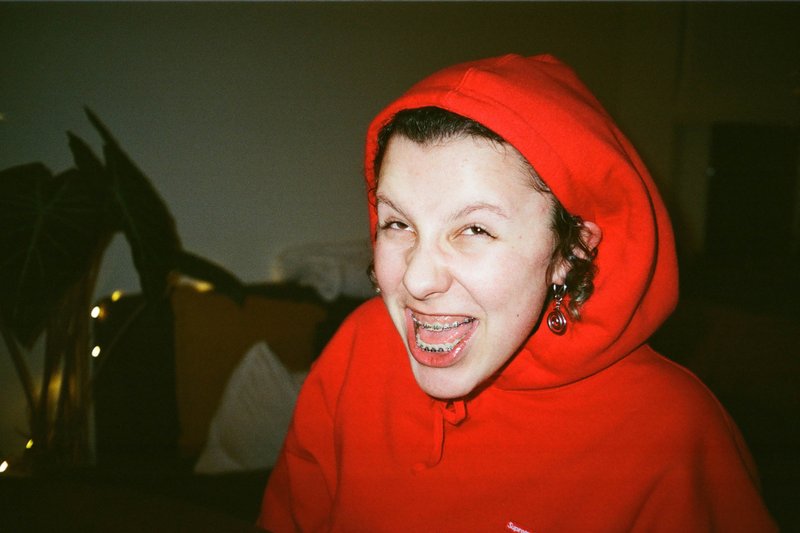 The image features a young person with a striking red hoodie. They are captured from a close distance, filling most of the frame. The individual appears to be indoors as there's a plant and a wall in the background, suggesting a home setting. The person is making a playful face towards the camera, possibly posing for the photo. The image has a darker tone, with a more muted color palette apart from the red hoodie that stands out prominently.