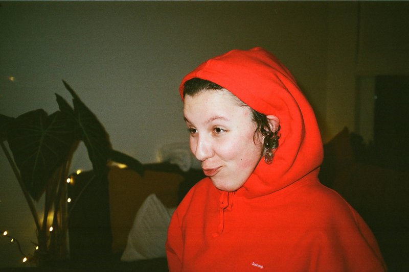 The image captures a young individual, possibly a child or teenager, wearing a red hoodie with a white emblem on the front. The person is making a playful expression with their mouth open and has a serious expression on their face. They are looking off to the side. The background is indistinct but appears to be an interior setting with some sort of lighting or fixture visible.