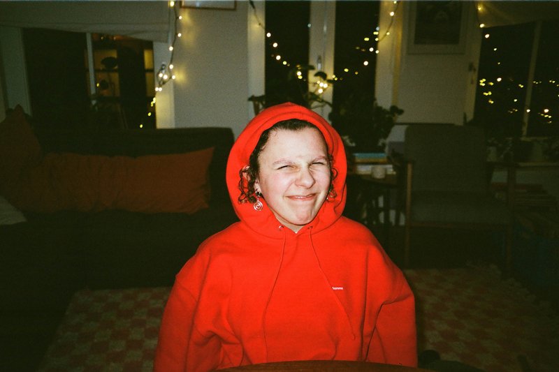 The image shows a young individual wearing a red hoodie with their face partially obscured by it. They are seated indoors with a cozy ambiance, indicated by the warm lighting and the glimpse of Christmas lights in the background. The individual appears to be in a casual setting, possibly a living room or similar domestic space, as suggested by the presence of a couch and other household items that are not clearly visible due to the low-resolution image. The person's expression is one of slight discomfort, perhaps indicated by the hoodie being bunched up near their face.