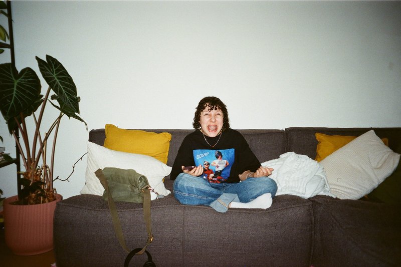 In the image, there is a woman sitting on a couch. She appears to be engaged with something off-camera, her hand is raised up as if she's gesturing or interacting with someone or something out of frame. The couch she's seated on has a patterned pillow. Behind her, there are two potted plants placed on a surface that seems like it could be a window sill or a tabletop. On the floor next to the couch, there is a backpack or bag with a visible logo or design.