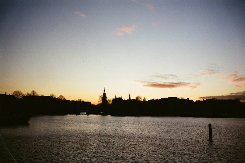 The image captures a serene sunset scene with a calm body of water reflecting the sky. In the distance, silhouettes of buildings can be seen on the opposite shore. The color palette consists of deep blues, purples, and warm yellows from the setting sun.