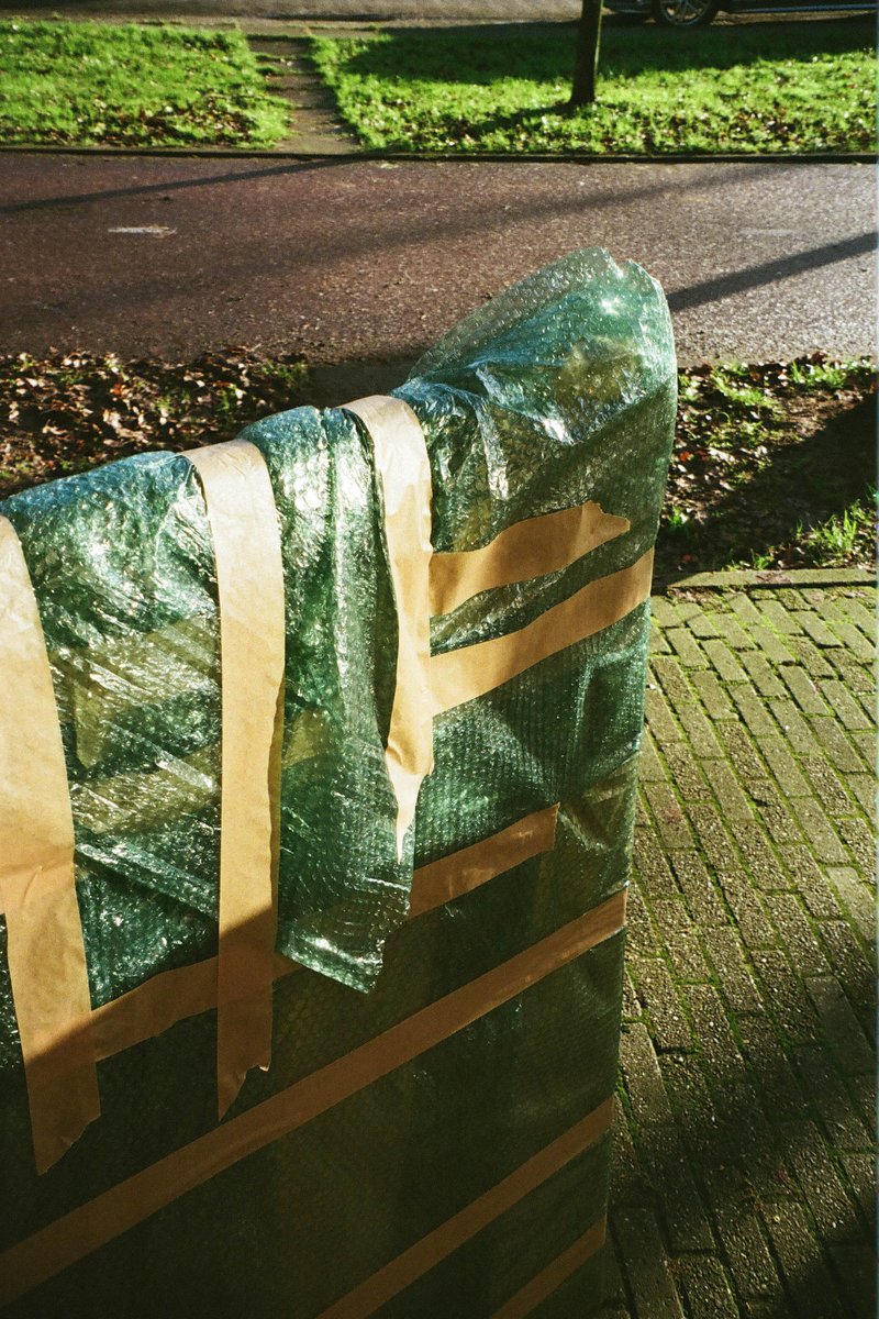 The image features a large, unwrapped object placed against a brick wall outside. It is covered with a layer of green plastic sheeting. The item appears to be wrapped up for protection or storage. The background indicates an outdoor setting with a paved ground and some greenery visible in the distance.