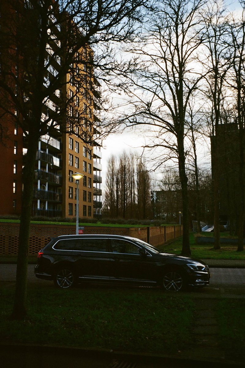 The image shows an urban scene featuring a residential area during daylight. The primary focus is on two cars parked on a street in front of apartment buildings, which are likely high-rise structures given their height and the presence of balconies. The sky appears overcast with no visible clouds. There's a sense of orderliness to the image, as evidenced by the neatly kept area and the structured layout of the buildings. The lighting suggests it might be an early morning or late afternoon due to the soft and diffused light, casting long shadows on the ground.