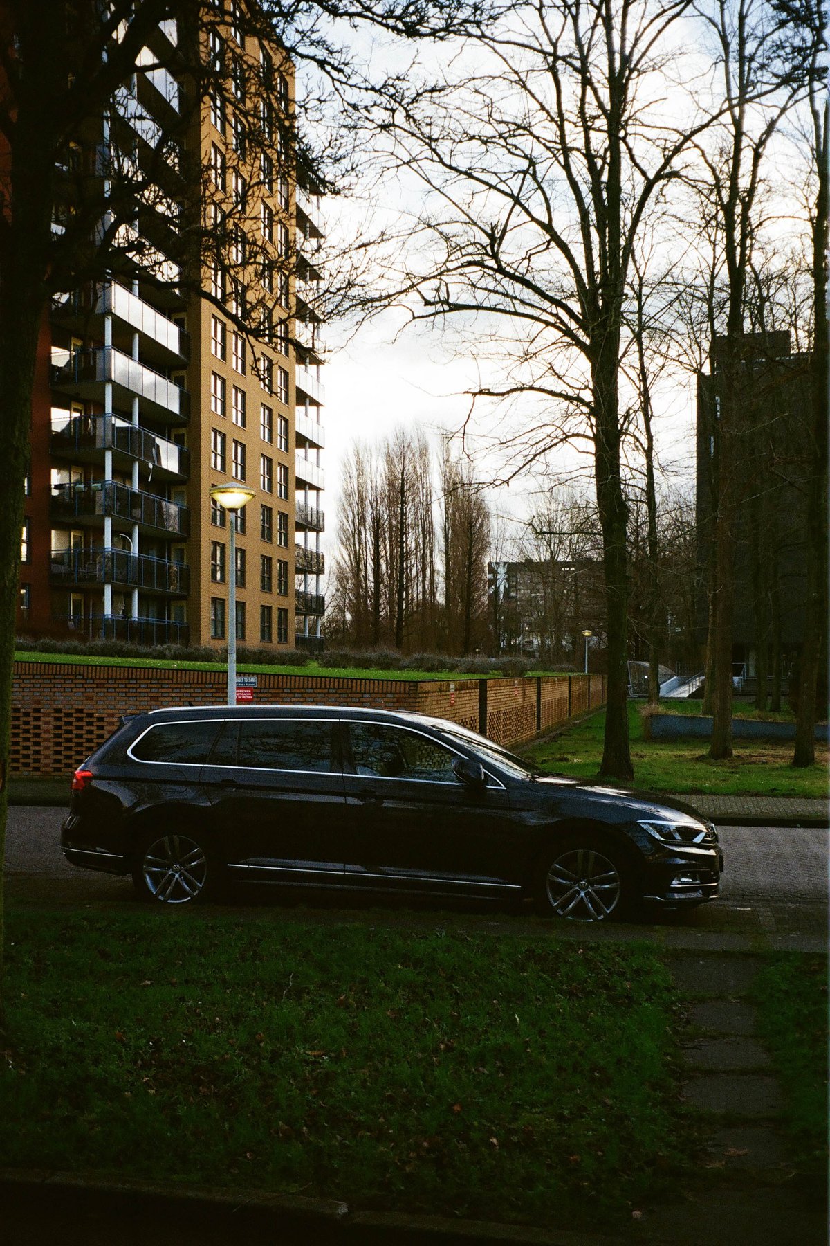  The image depicts a residential area with a dark SUV parked near a sidewalk, adjacent to a fence and a few trees. In the background, there is a building and a sky that appears to be overcast. The colors are muted, with the greys of the car, trees, and the overcast sky being dominant. The photo has an artistic filter applied, giving it a slightly abstracted quality. 