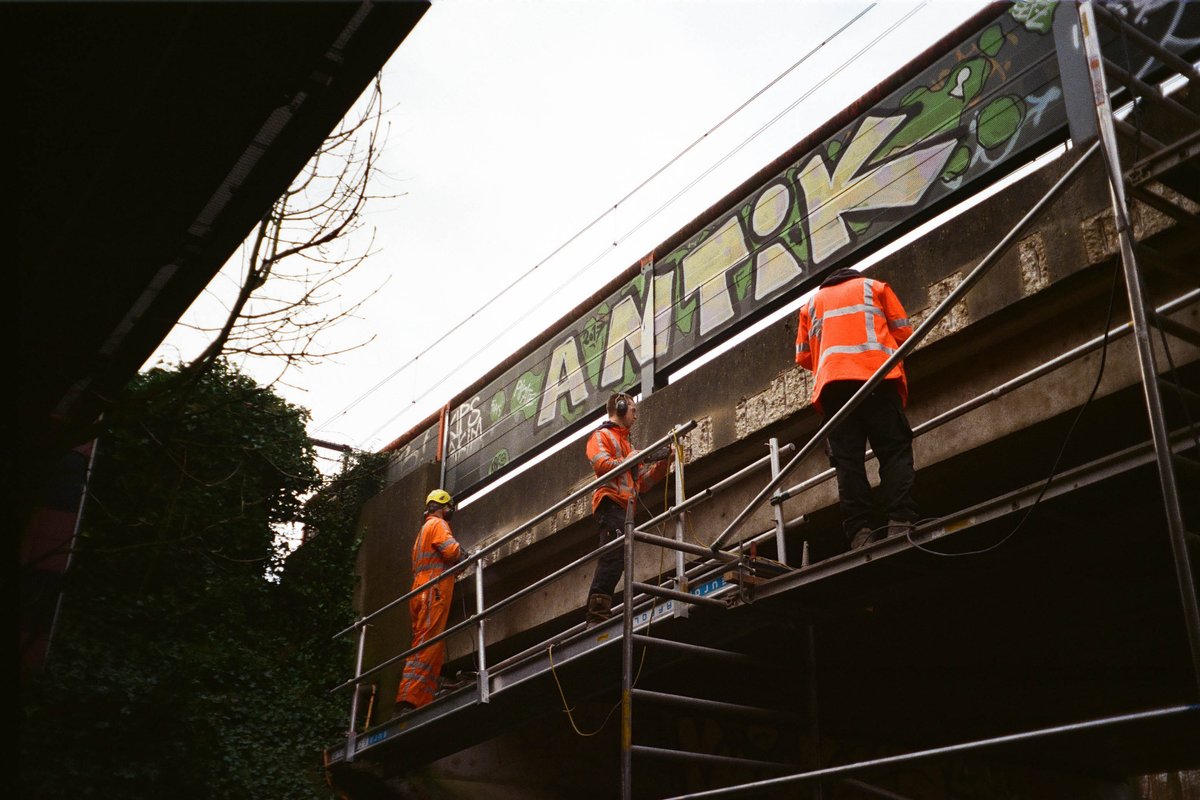 The image portrays a scene of urban development or maintenance. There's a construction site with two workers visible. They are engaged in activities related to the work being done on a wall or structure nearby, possibly installing or repairing it. One worker is seen holding up a sign that reads "YOU ARE WATCHING THE NEWS," which adds an element of humor and commentary to the scene. The workers are dressed in standard construction attire, suggesting they might be professionals at their craft. There is also a bridge with graffiti on it in the background. The colors in the image are quite muted, with shades of grey, green, and orange dominating the visual palette. 