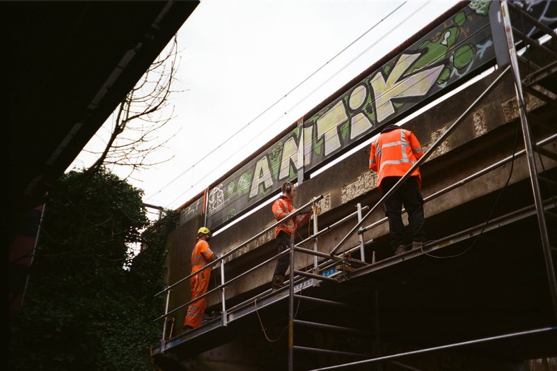 The image portrays a scene of urban development or maintenance. There's a construction site with two workers visible. They are engaged in activities related to the work being done on a wall or structure nearby, possibly installing or repairing it. One worker is seen holding up a sign that reads "YOU ARE WATCHING THE NEWS," which adds an element of humor and commentary to the scene. The workers are dressed in standard construction attire, suggesting they might be professionals at their craft. There is also a bridge with graffiti on it in the background. The colors in the image are quite muted, with shades of grey, green, and orange dominating the visual palette.