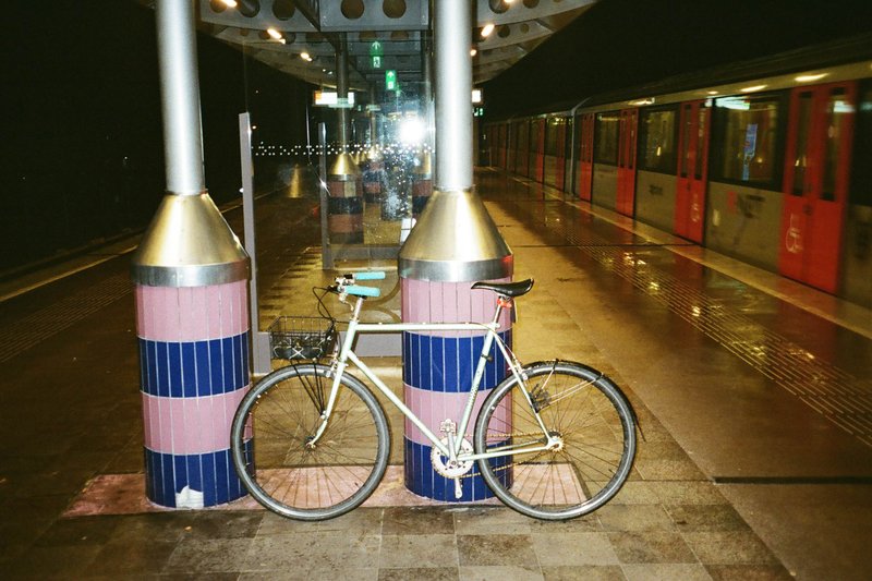 This image captures a peaceful night scene at a train station. There is a bicycle parked by a blue and pink pillar under a large light. In the background, there are two trains, one partially visible on the left side of the image and another fully visible on the right, waiting for passengers. The platform appears to be wet from recent rain, adding a reflective quality to the scene.