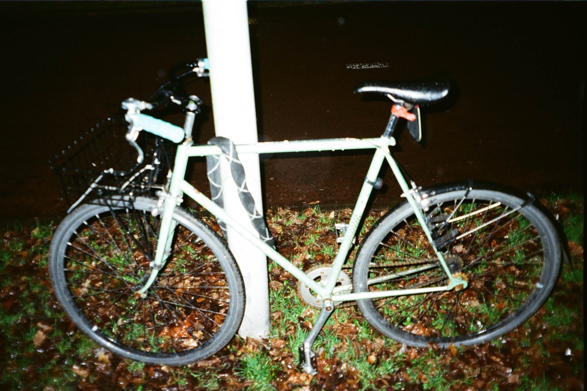  The image presents an old-fashioned bicycle parked next to a streetlamp post during nighttime. The bike is leaning against the pole, suggesting it's momentarily unattended or possibly locked there for security. It features a prominent bell and a basket on the handlebars, hinting at its utilitarian purpose. 