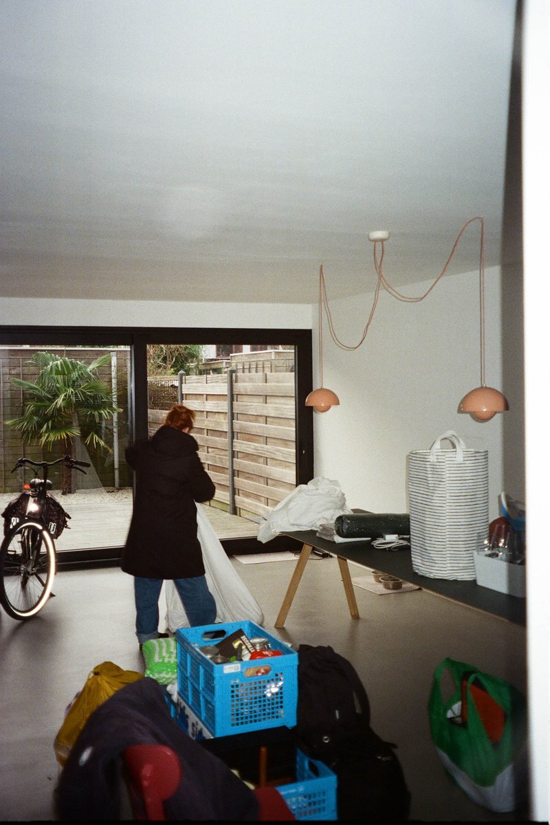 This is an indoor scene with a person standing near a table cluttered with various items. There's a bicycle parked inside, suggesting that outdoor activities are nearby. The room appears to be a storage area or a workspace, given the presence of packing materials and boxes in the background. The wall decoration includes a series of vertical stripes, which might be an artistic choice for wallpaper or a design feature. The lighting is bright, suggesting it's daytime.