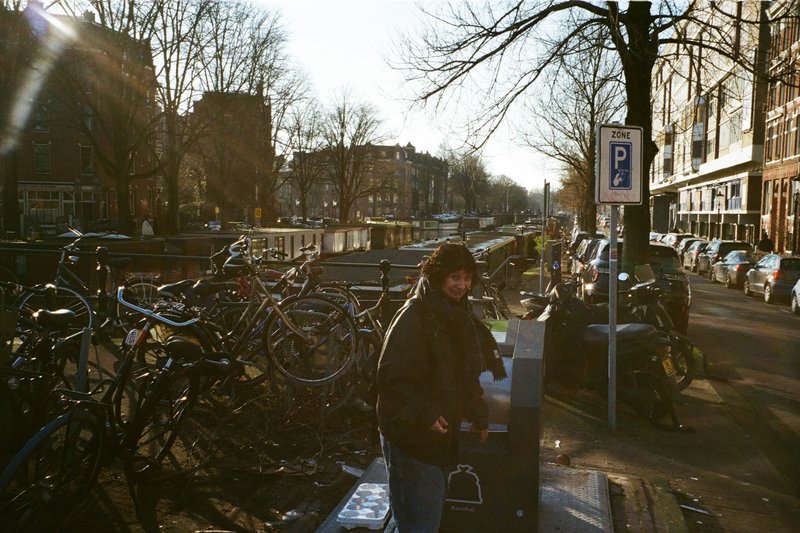 The image depicts an urban scene with a man standing by a bicycle rack, which is situated near parked bicycles. The bicycles are attached to a metal frame, likely providing secure parking for cyclists. In the background, there's a city street lined with parked cars and a building that features a clock tower. A person wearing sunglasses can be seen walking down the sidewalk towards a bench under a tree. A blue sky with some clouds is above the scene, suggesting a clear day.