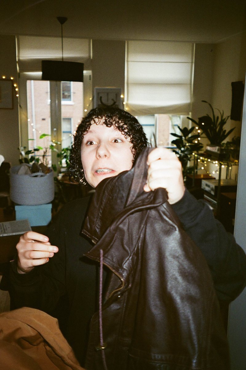 The image is of a person standing indoors, holding up what appears to be a garment or jacket. This individual seems to be showing off the item to the camera. The setting suggests a casual indoor environment with furniture visible in the background. There are no significant landmarks or recognizable symbols that can give more specific context about the location.