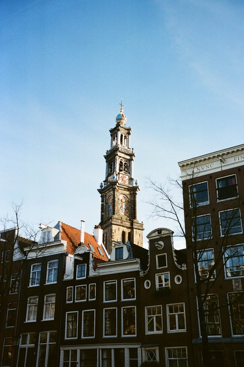 The image features a tall clock tower with a bell at its pinnacle, prominently situated amidst other buildings in an urban landscape. The sky is clear and blue, suggesting it might be daytime. The photo seems to have been taken from street level, looking up towards the sky and architecture.