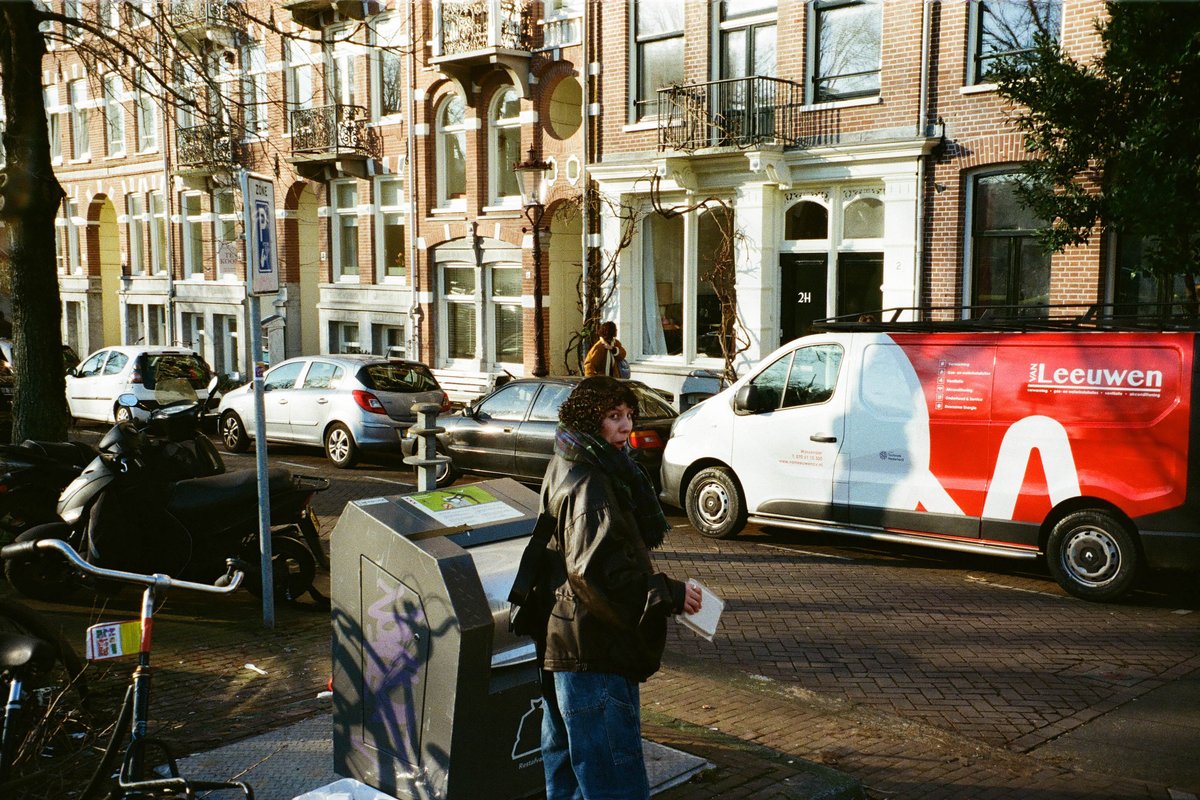  The image shows an urban street scene with a tall white building featuring large windows and balconies. In front of the building is parked a vehicle with a visible advertisement for "LEJVEN" on its side, depicting a woman's face. There are people walking on the sidewalk, one person is closer to the camera, while another person is farther away. The street is lined with parked cars, and there is a bicycle leaning against something out of frame. 