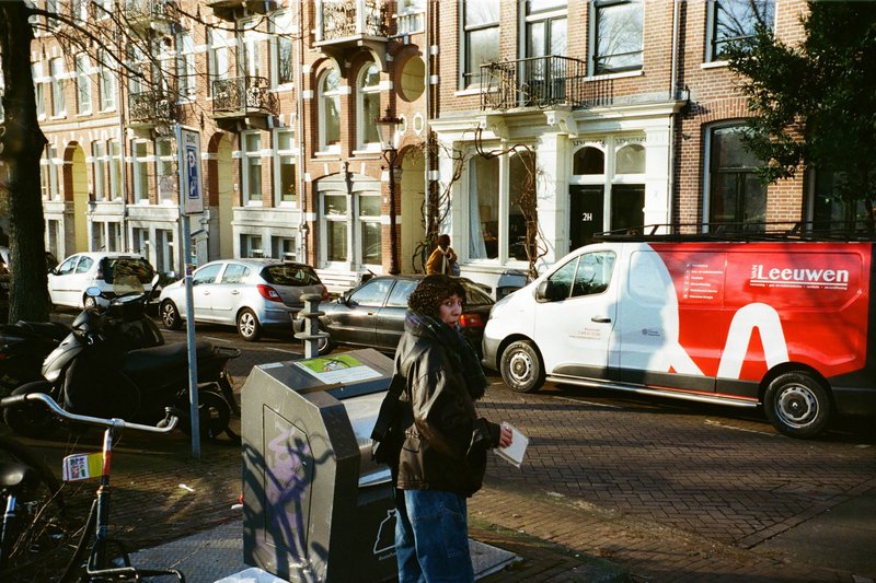 The image shows an urban street scene with a tall white building featuring large windows and balconies. In front of the building is parked a vehicle with a visible advertisement for "LEJVEN" on its side, depicting a woman's face. There are people walking on the sidewalk, one person is closer to the camera, while another person is farther away. The street is lined with parked cars, and there is a bicycle leaning against something out of frame.