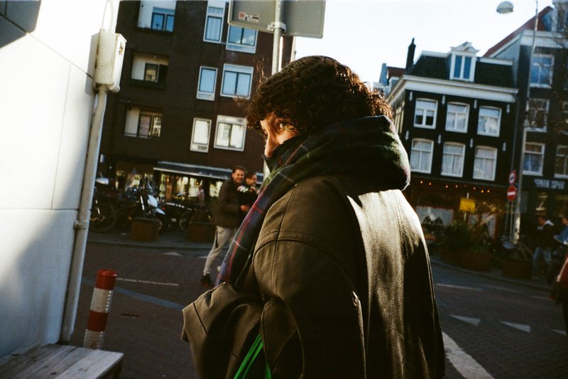 The image depicts a person standing on a city street corner during what appears to be daytime. The individual is looking towards the right of the frame, with their back to the camera. They are dressed in a dark coat, and their hair is brown. Behind the person, there is a signpost and some buildings visible in the background. There are no other people immediately apparent nearby, just the main subject standing alone. The lighting suggests it's daytime, possibly late afternoon or early morning due to the diffuse light.