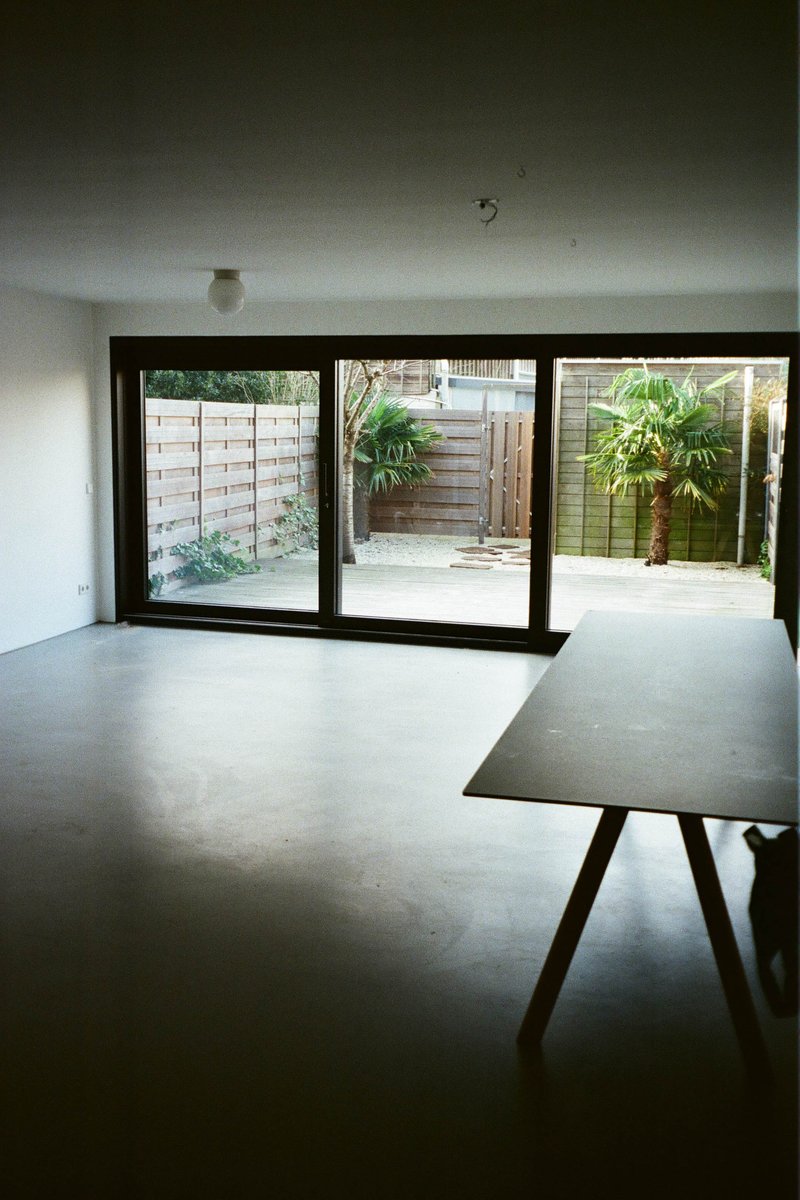 The image depicts an interior space, likely a room with a view of a yard through windows, which are visible but not the primary focus. There's a dining table in the foreground on the right side. The flooring is not distinctly visible, and no significant text or distinguishing features that could provide additional context can be discerned.